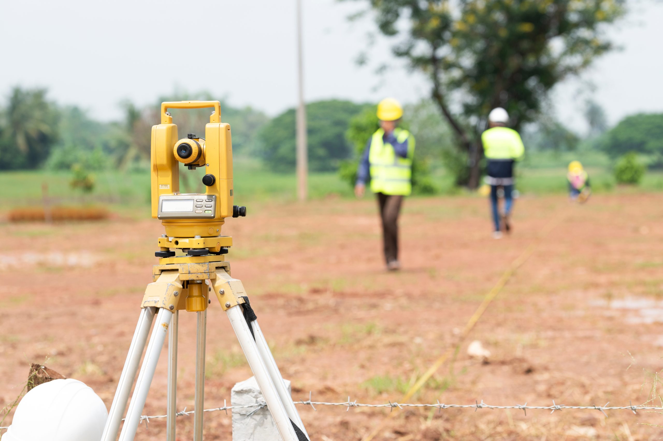 A yellow surveying instrument is positioned on a tripod in the foreground, with two individuals in safety vests walking in the background on a cleared, brown field. Trees and a cloudy sky are visible in the distance