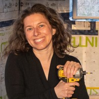 A woman with curly hair smiles while holding a power drill. She is wearing a black top and is in a construction or renovation setting, with building materials visible in the background