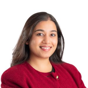 A woman with long, dark hair smiles while wearing a red blazer. She has a friendly expression and is positioned against a white background