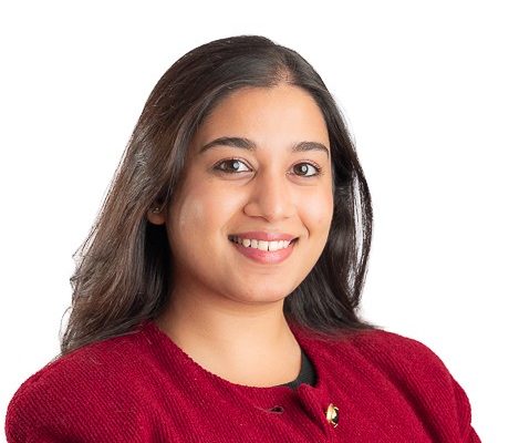 A woman with long, dark hair smiles while wearing a red blazer. She has a friendly expression and is positioned against a white background