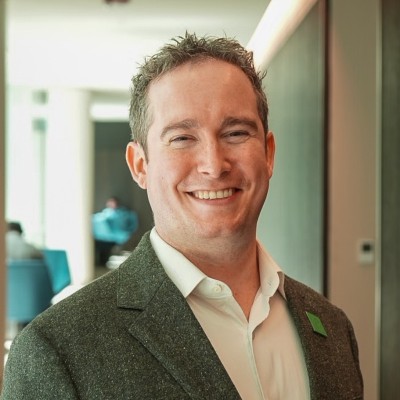 A man with short, curly hair smiles while wearing a gray blazer over a white shirt. The background features a modern indoor setting with blurred figures and furniture