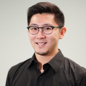 A young man with short black hair and glasses smiles at the camera, wearing a black button-up shirt. The background is a light gray color