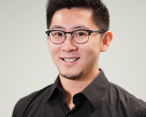 A young man with short black hair and glasses smiles at the camera, wearing a black button-up shirt. The background is a light gray color