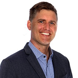A man with short, styled hair and a friendly smile is wearing a dark blazer over a light blue shirt. He is positioned against a plain white background