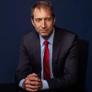 A man in a dark suit and red tie sits at a table, resting his hands together. He has short, light brown hair and a serious expression, with a dark blue background behind him