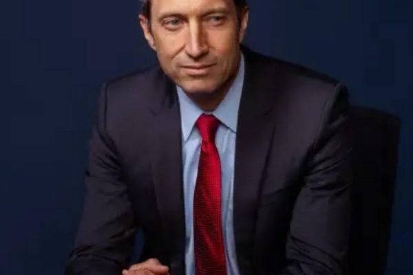 A man in a dark suit and red tie sits at a table, resting his hands together. He has short, light brown hair and a serious expression, with a dark blue background behind him