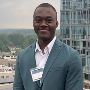 A man stands outdoors in front of a modern building, wearing a light blue blazer and a white shirt. He has a name tag attached to his blazer and is smiling at the camera. The background features a cityscape with greenery and other buildings
