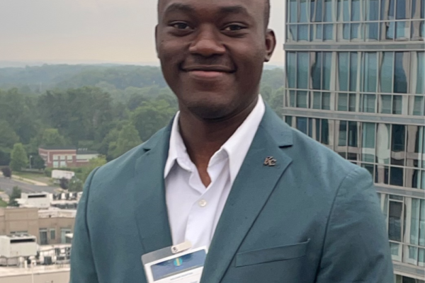 A man stands outdoors in front of a modern building, wearing a light blue blazer and a white shirt. He has a name tag attached to his blazer and is smiling at the camera. The background features a cityscape with greenery and other buildings