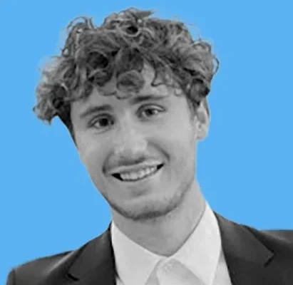 A young man with curly hair smiles at the camera, wearing a suit and a collared shirt. The background is a solid light blue color