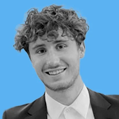A young man with curly hair smiles at the camera, wearing a suit and a collared shirt. The background is a solid light blue color