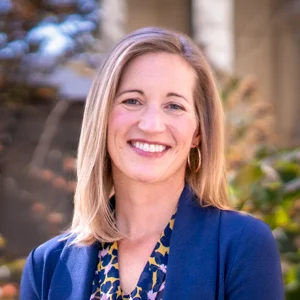 A woman with shoulder-length blonde hair smiles while wearing a blue blazer over a patterned blouse. She stands outdoors with greenery in the background