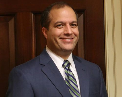 A man stands smiling in front of a wooden door. He is wearing a dark suit with a light blue shirt and a striped tie in shades of blue and green