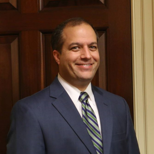 A man stands smiling in front of a wooden door. He is wearing a dark suit with a light blue shirt and a striped tie in shades of blue and green