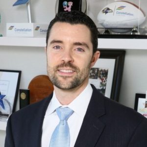 A man with dark hair and a beard is wearing a suit and light blue tie. He is smiling and standing in front of a shelf displaying various awards and memorabilia, including a football and a star-shaped trophy