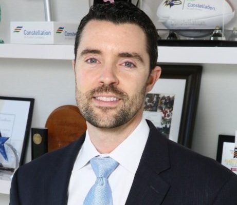 A man with dark hair and a beard is wearing a suit and light blue tie. He is smiling and standing in front of a shelf displaying various awards and memorabilia, including a football and a star-shaped trophy