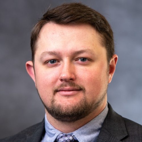 A man with short brown hair and a beard is wearing a gray suit and a light blue shirt with a tie. He is looking directly at the camera with a neutral expression. The background is a blurred gray