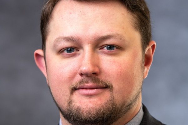 A man with short brown hair and a beard is wearing a gray suit and a light blue shirt with a tie. He is looking directly at the camera with a neutral expression. The background is a blurred gray