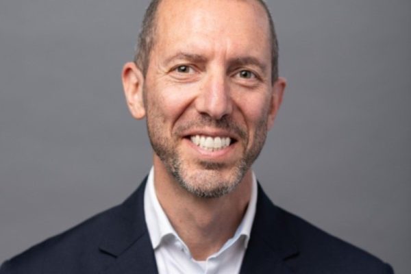 A man with short, light brown hair and a beard smiles at the camera. He is wearing a dark blazer over a white shirt. The background is a neutral gray
