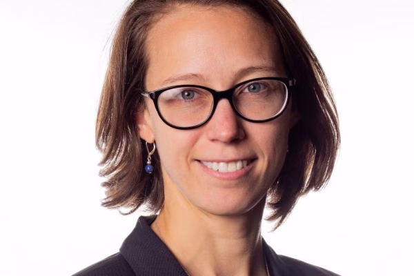 A woman with shoulder-length brown hair and glasses smiles at the camera. She is wearing a black blazer and has a small earring. The background is light and neutral
