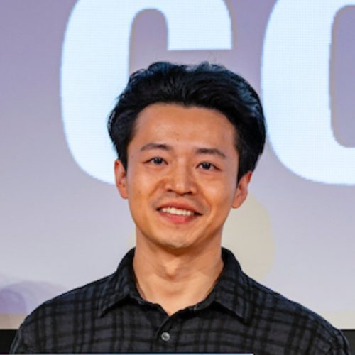 A young man with dark hair and a friendly smile stands in front of a large screen displaying the word "GO." He is wearing a black plaid shirt