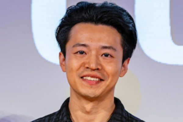 A young man with dark hair and a friendly smile stands in front of a large screen displaying the word "GO." He is wearing a black plaid shirt