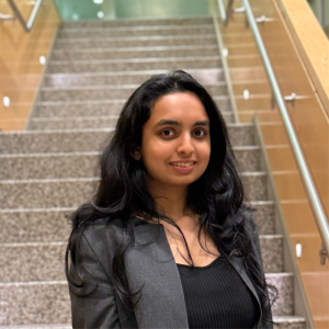 A young woman with long black hair stands on a staircase. She is wearing a black top and a gray blazer, smiling at the camera. The staircase has a modern design with a light-colored railing and stone steps
