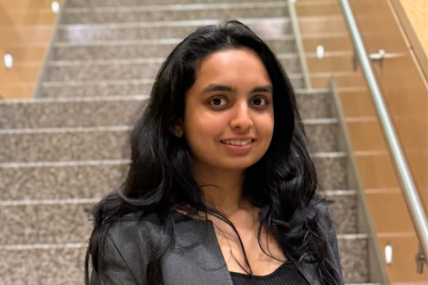 A young woman with long black hair stands on a staircase. She is wearing a black top and a gray blazer, smiling at the camera. The staircase has a modern design with a light-colored railing and stone steps