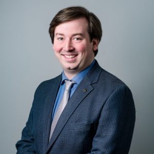 A man with medium-length brown hair smiles while wearing a dark blazer over a light blue shirt and a tie. He stands against a plain gray background
