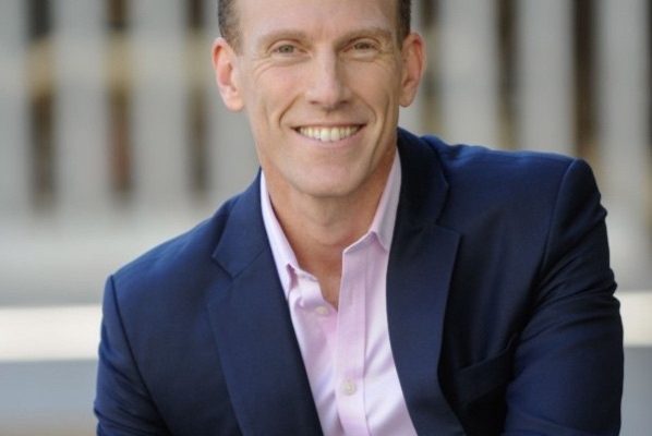 A man with short, light brown hair is smiling while seated. He is wearing a dark blue suit jacket over a light pink shirt. The background features blurred architectural elements