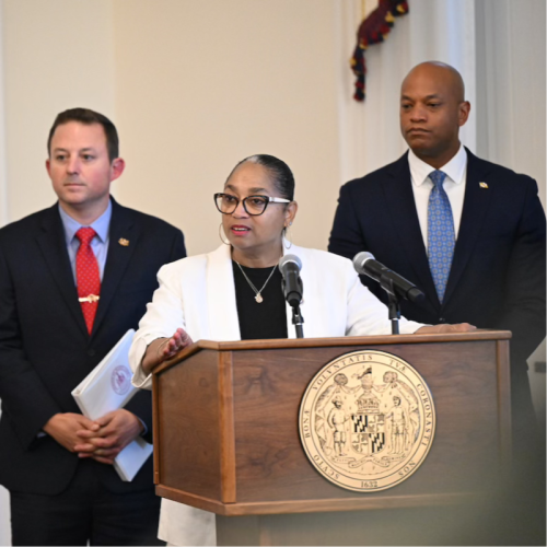A woman wearing glasses and a white blazer stands at a podium with a state seal, speaking into a microphone. Two men in suits stand behind her, one holding a folder. The setting appears to be an official event or press conference