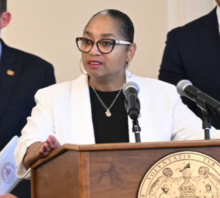 A woman wearing glasses and a white blazer stands at a podium with a state seal, speaking into a microphone. Two men in suits stand behind her, one holding a folder. The setting appears to be an official event or press conference