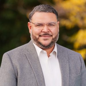 A man with short, curly hair and a beard is smiling while wearing glasses. He is dressed in a light-colored shirt and a gray blazer. The background features blurred greenery and autumn foliage