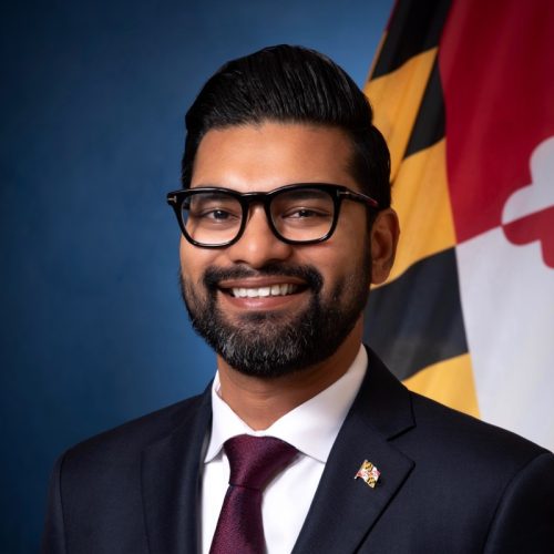 A man with a beard and glasses smiles confidently while wearing a dark suit and a maroon tie. He is positioned in front of a blue background and a Maryland state flag