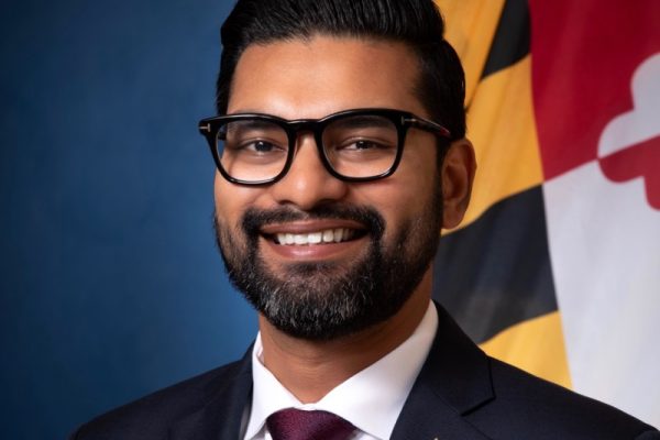 A man with a beard and glasses smiles confidently while wearing a dark suit and a maroon tie. He is positioned in front of a blue background and a Maryland state flag