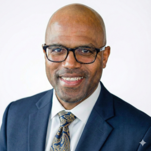 A smiling man with a bald head and glasses is wearing a dark suit and a patterned tie. He has a friendly expression and is positioned against a light background