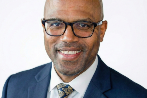 A smiling man with a bald head and glasses is wearing a dark suit and a patterned tie. He has a friendly expression and is positioned against a light background
