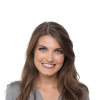 A woman with long, wavy brown hair and a bright smile is wearing a gray blouse. She is facing the camera against a plain white background