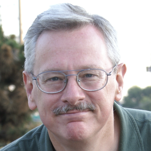 A middle-aged man with gray hair and glasses smiles at the camera. He has a mustache and is wearing a dark green shirt. The background features blurred greenery and a hint of a road
