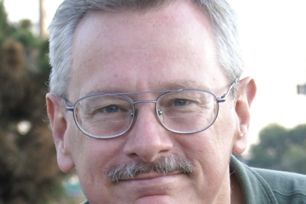 A middle-aged man with gray hair and glasses smiles at the camera. He has a mustache and is wearing a dark green shirt. The background features blurred greenery and a hint of a road