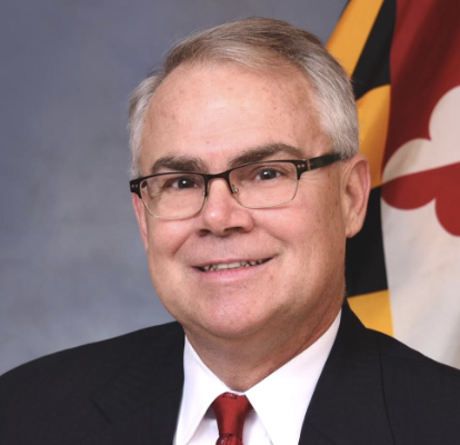 A middle-aged man with gray hair and glasses smiles while wearing a suit and tie. He is positioned in front of a Maryland state flag