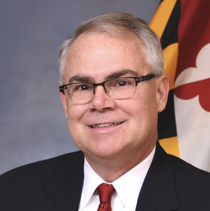 A middle-aged man with gray hair and glasses smiles while wearing a suit and tie. He is positioned in front of a Maryland state flag