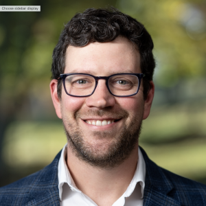 A man with curly dark hair and glasses smiles at the camera. He is wearing a white shirt and a blue blazer. The background features blurred greenery, suggesting an outdoor setting