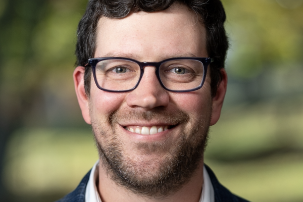 A man with curly dark hair and glasses smiles at the camera. He is wearing a white shirt and a blue blazer. The background features blurred greenery, suggesting an outdoor setting