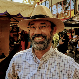 A man with a beard and glasses smiles while wearing a straw hat and a checkered shirt. He is standing outdoors, with people and market stalls visible in the background