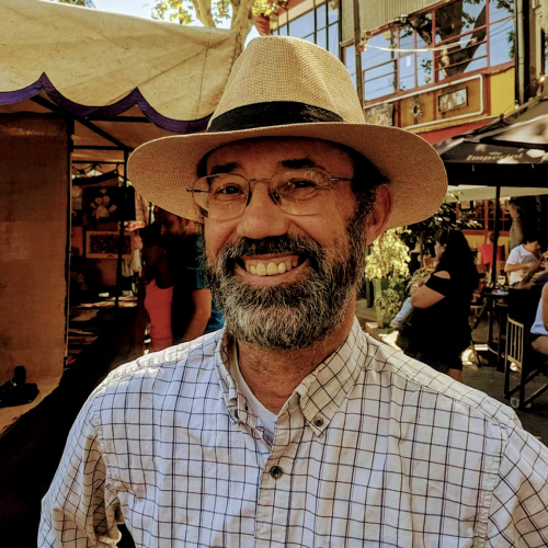 A man with a beard and glasses smiles while wearing a straw hat and a checkered shirt. He is standing outdoors, with people and market stalls visible in the background