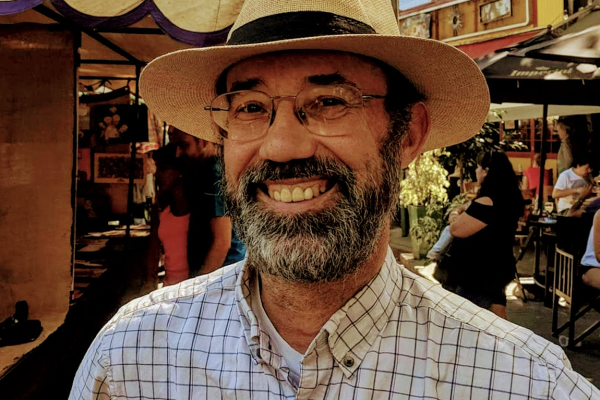 A man with a beard and glasses smiles while wearing a straw hat and a checkered shirt. He is standing outdoors, with people and market stalls visible in the background