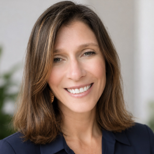 A woman with shoulder-length brown hair and a warm smile is looking directly at the camera. She is wearing a dark blue blouse and has earrings. The background features soft, blurred greenery