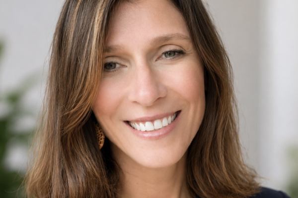 A woman with shoulder-length brown hair and a warm smile is looking directly at the camera. She is wearing a dark blue blouse and has earrings. The background features soft, blurred greenery
