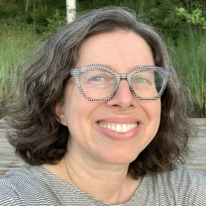 A person with curly, shoulder-length hair smiles at the camera, wearing clear, patterned glasses and a striped shirt. The background features greenery and a wooden surface