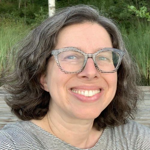 A person with curly, shoulder-length hair smiles at the camera, wearing clear, patterned glasses and a striped shirt. The background features greenery and a wooden surface